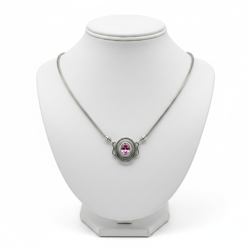 A silver-toned chain necklace with an oval pendant featuring a pink gemstone, displayed on a white jewelry bust against a white background.